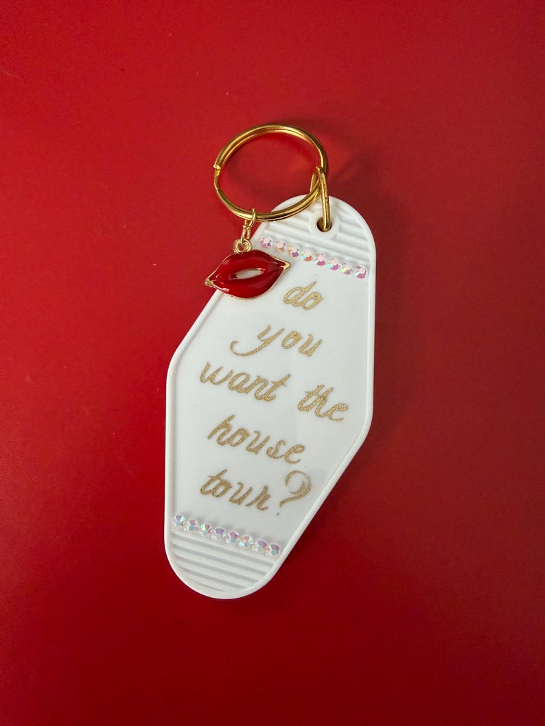 May include: White key tag with gold text that reads "do you want the house tour?" A red lip charm and clear rhinestones adorn the tag, which hangs from a gold key ring. The background is a solid red.