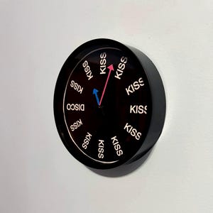 May include: Black wall clock with the word "KISS" in white lettering around the clock face. The clock hands are pink and blue. The word "DISCO" is also visible on the clock face.