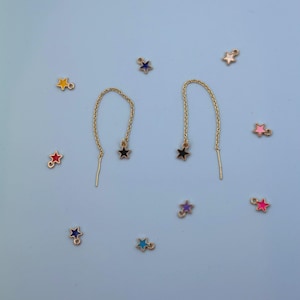 May include: Gold-tone chain earrings with a black star charm, and a collection of colorful star charms scattered on a light blue background. The earrings have a delicate chain design, and the star charms are in various colors.