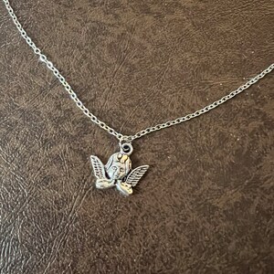以下が含まれることがあります： 小さな天使のペンダントが付いたシルバーチェーンネックレス。天使は翼があり、下を見ている。