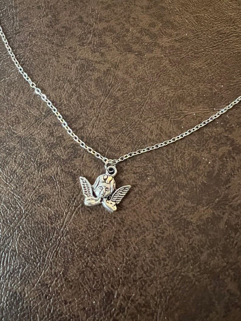以下が含まれることがあります： 小さな天使のペンダントが付いたシルバーチェーンネックレス。天使は翼があり、下を見ている。