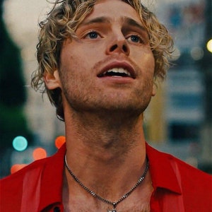 以下が含まれることがあります： 金色の巻き毛の男性が赤いシャツを着て、小さな天使のペンダントが付いた銀のチェーンネックレスをしています。彼は上を見上げて微笑んでいます。