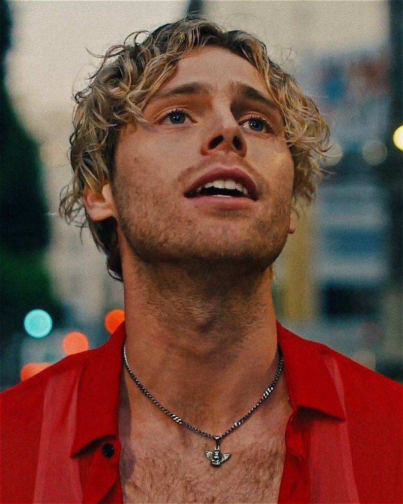 以下が含まれることがあります： 金色の巻き毛の男性が赤いシャツを着て、小さな天使のペンダントが付いた銀のチェーンネックレスをしています。彼は上を見上げて微笑んでいます。