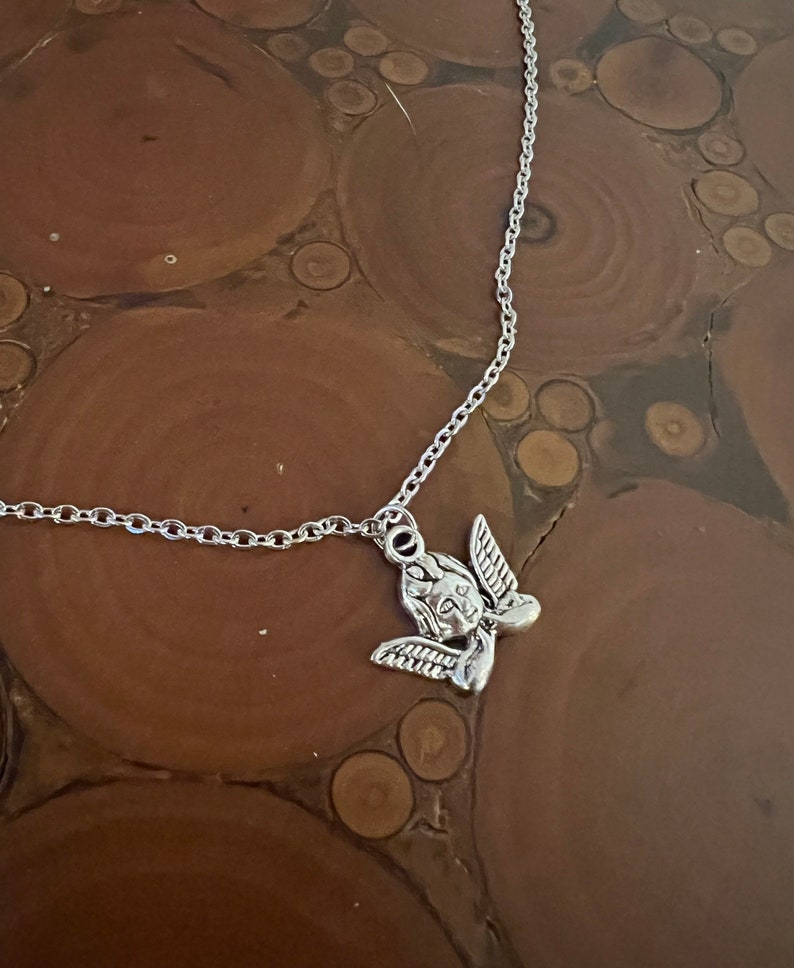 以下が含まれることがあります： 小さな天使のペンダントが付いたシルバーチェーンネックレス。天使は前を向いて翼を広げています。