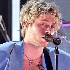 以下が含まれることがあります： 金色の巻き毛の男性が薄い青色のブレザーとペンダント付きの銀色のネックレスを着ています。彼はマイクに向かって歌っています。