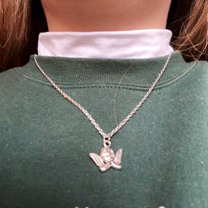 以下が含まれることがあります： 小さな天使の羽根のペンダントが付いたシルバーチェーンネックレス。ネックレスは白いタートルネックと緑色のスウェットシャツの上に着用されています。