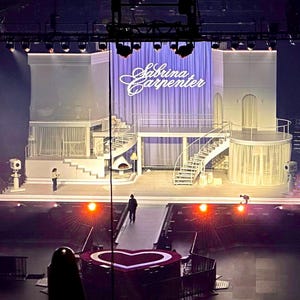 May include: A stage set with multiple levels, spiral staircases, and a heart-shaped platform. The backdrop features a purple curtain with the name "Sabrina Carpenter" in cursive. The lighting is dramatic, with spotlights and stage lights.