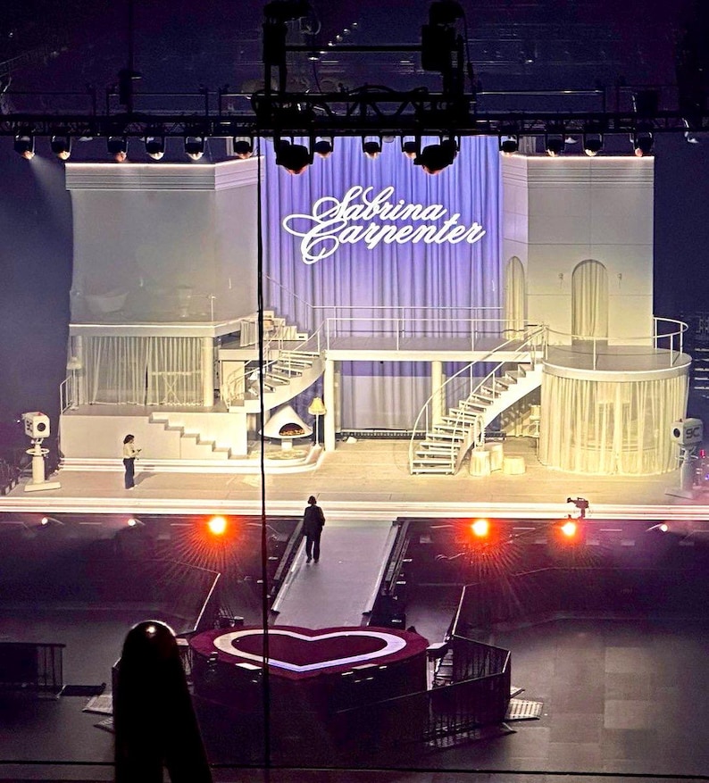 May include: A stage set with multiple levels, spiral staircases, and a heart-shaped platform. The backdrop features a purple curtain with the name "Sabrina Carpenter" in cursive. The lighting is dramatic, with spotlights and stage lights.
