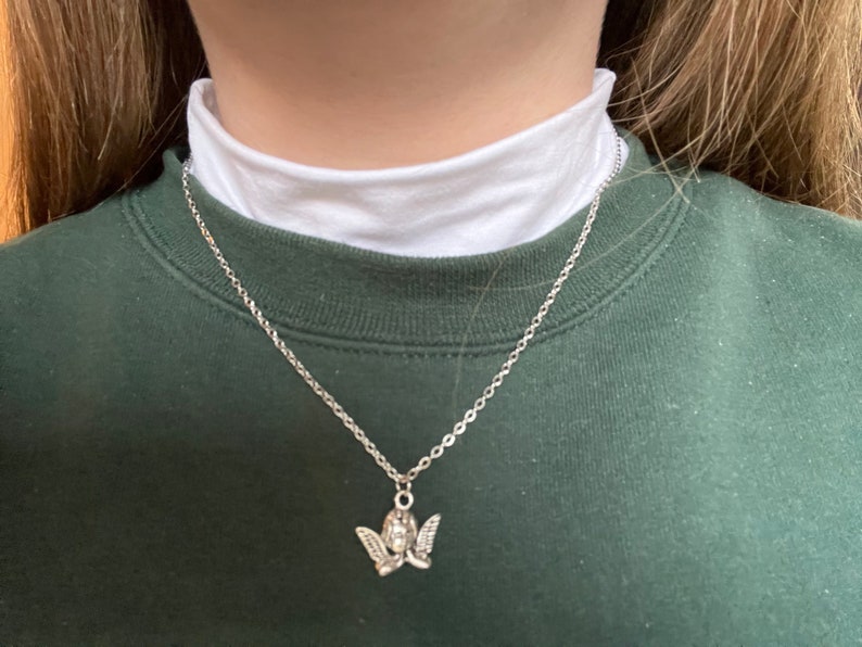 以下が含まれることがあります： 天使のペンダントが付いたシルバーチェーンネックレス。天使は翼とハローで描かれています。