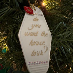 May include: A white motel-style keychain ornament with gold lettering that reads "do you want the house tour?" A red lip charm and small crystals adorn the top and bottom. The ornament hangs from a gold ring.