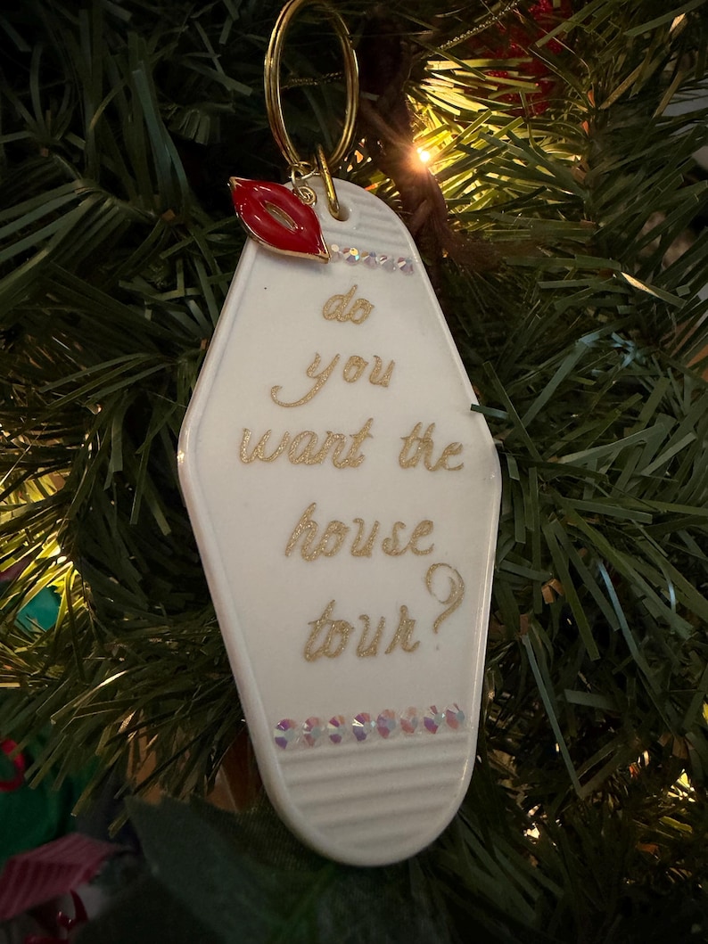 May include: A white motel-style keychain ornament with gold lettering that reads "do you want the house tour?" A red lip charm and small crystals adorn the top and bottom. The ornament hangs from a gold ring.