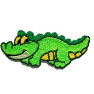 Può includere: Toppa ricamata verde di un alligatore dei cartoni animati con accenti gialli.
