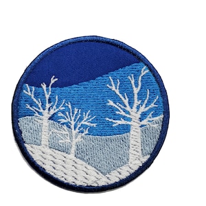 May include: Embroidered patch with a winter scene. The patch is circular and features three white trees against a blue sky with a white snowy ground.
