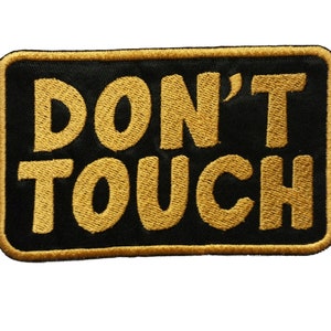 May include: A rectangular black patch with the words "DON'T TOUCH" embroidered in gold thread. The patch has a gold border and is set against a white background.