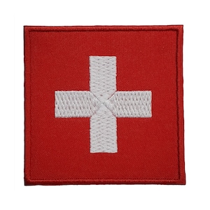 Puede incluir: Un parche cuadrado rojo con una cruz blanca bordada en el centro. El parche está bordeado con costuras blancas.