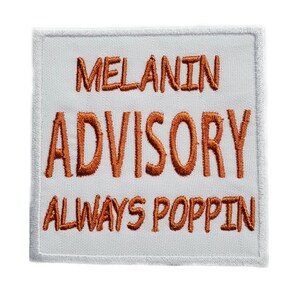 May include: White fabric patch with orange embroidered text that reads "MELANIN ADVISORY ALWAYS POPPIN"