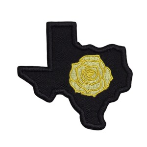 Puede incluir: Un parche bordado con la forma del estado de Texas, que presenta un fondo negro con un diseño de rosa amarilla centrado en el medio. El parche tiene un borde grueso cosido en negro que delinea toda la forma del estado y la rosa.