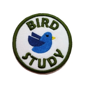 May include: Embroidered patch with a blue bird illustration and the words "BIRD STUDY" in green. The bird has a yellow beak and is set against a white background, framed by a green border. Ideal for jackets, bags, or other fabric items.