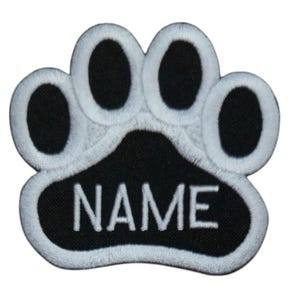 May include: Embroidered paw print patch in black and white. The patch features a black paw print with white stitching around the edges. The word "NAME" is embroidered in white letters on the black center.