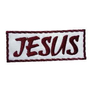 Pode incluir: Um remendo branco com uma borda bordada marrom e a palavra "JESUS" em letras marrons.