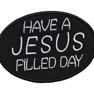 May include: Black oval patch with white embroidered text that reads "HAVE A JESUS FILLED DAY". The patch has a black border and is likely designed to be sewn or ironed onto clothing or accessories.