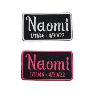 May include: Two rectangular embroidered patches. The top patch has a black background with white text that reads "Naomi" and the dates "1/11/46 - 4/30/22". The bottom patch has a black background with pink text that reads "Naomi" and the dates "1/11/46 - 4/30/22".