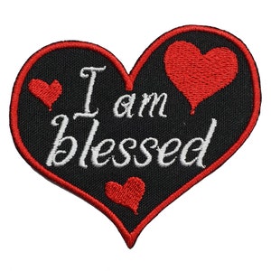 May include: Black heart-shaped patch with red stitching and three red hearts. The text "I am blessed" is embroidered in white on the patch.