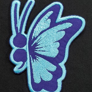 May include: Embroidered butterfly patch in shades of blue and purple. The butterfly design features a detailed wing pattern and a light blue outline. The patch is likely for clothing or accessories.