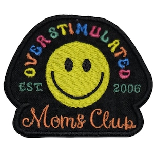 May include: A black embroidered patch shaped like a mushroom cap featuring a yellow smiley face in the center. The text OVERSTIMULATED is arched in multicolored letters above the face, with EST. 2006 and the script text Moms Club below.
