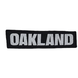 Op de afbeelding: Een rechthoekige geborduurde patch met een zwarte achtergrond en het woord OAKLAND gestikt in dikke, lichtgrijze hoofdletters. De patch heeft een strakke, gestikte rand en een platte, horizontale oriëntatie met een duidelijk schreefloos lettertype.