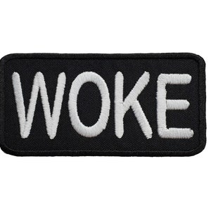 May include: Black rectangular patch with the white embroidered word "WOKE". The patch has rounded corners and a black border. The white text is in a bold, sans-serif font.