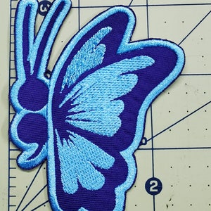 May include: Embroidered butterfly patch in shades of blue. The patch features a dark blue outline and a lighter blue fill, with detailed stitching. The butterfly design is symmetrical and suitable for clothing or accessories.