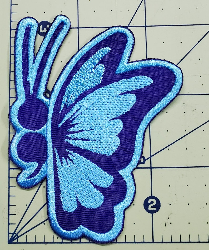 May include: Embroidered butterfly patch in shades of blue. The patch features a dark blue outline and a lighter blue fill, with detailed stitching. The butterfly design is symmetrical and suitable for clothing or accessories.