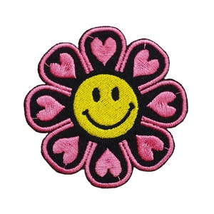 May include: A pink and black embroidered patch with a smiley face in the centre and hearts around the edge.
