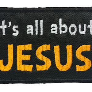 Könnte beinhalten: Schwarzer gestickter Aufnäher mit weißem Text, der "It's all about" lautet, und gelbem Text, der "JESUS" lautet.
