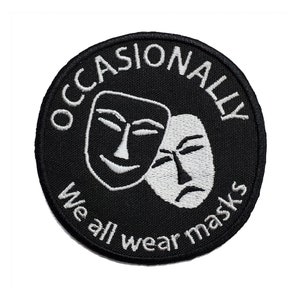 May include: A black and white embroidered patch with the text "Occasionally We All Wear Masks" in a circle around two white masks, one smiling and one frowning.