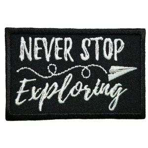 May include: Black rectangular patch with white embroidered text that reads "NEVER STOP EXPLORING". A white paper airplane graphic is to the right of the word "EXPLORING". The patch has a black border.