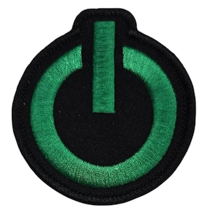 May include: A circular black patch featuring a green power symbol. The symbol consists of a vertical line above a semi-circle, all embroidered. The patch is edged with a black border.