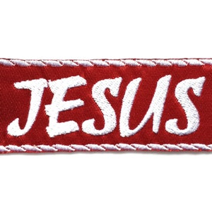 Pode incluir: Etiqueta bordada vermelha com costuras brancas. A etiqueta diz "JESUS" em uma fonte cursiva branca.