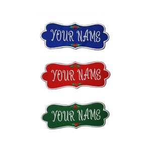 May include: Three embroidered name tags in blue, red, and green. Each tag has a white border and the words "YOUR NAME" in white script. Holly berry accents are above and below the text.