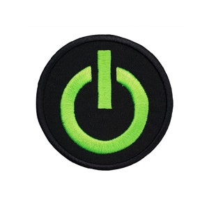 Può includere: Una toppa da stirare ricamata nera e verde con un simbolo di pulsante di accensione.