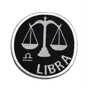 May include: A round, black embroidered patch featuring the Libra zodiac symbol in white. The scales of justice are centered above the word "LIBRA" in white, with the Libra symbol below the scales. The patch has a white border.