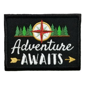 Op de afbeelding: Zwarte geborduurde patch met een kompas, groene bomen en de tekst "Adventure Awaits" met gouden pijlen die naar links en rechts wijzen.