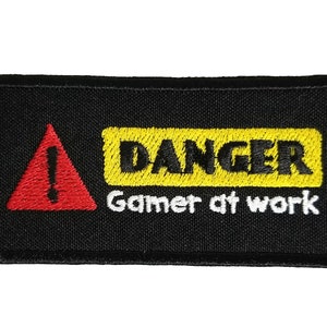 Pode incluir: Etiqueta bordada preta com um sinal de aviso triangular vermelho e o texto "DANGER Gamer at work" em amarelo e branco.