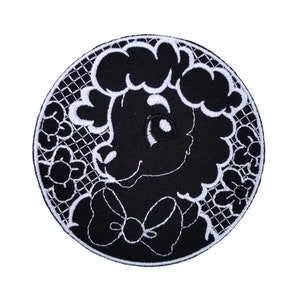 Puede incluir: Parche bordado en blanco y negro de una oveja de dibujos animados con un lazo. La oveja tiene una melena blanca rizada y un lazo blanco. El parche está rodeado por un diseño de encaje blanco.