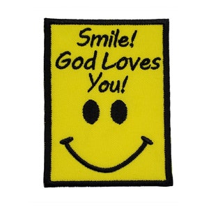 Puede incluir: Parche de tela amarillo con borde negro y una carita sonriente negra. El texto "Smile! God Loves You!" está bordado en negro.