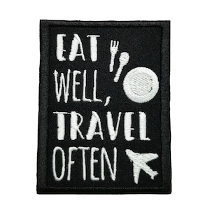 Op de afbeelding: Zwarte geborduurde patch met witte tekst die "Eat Well, Travel Often" luidt met een afbeelding van een vork, lepel, bord en vliegtuig.