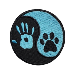May include: A circular embroidered patch featuring a yin and yang design. One side is black with a light blue handprint, and the other side is light blue with a black paw print. The entire patch is bordered by a thick black stitched edge.