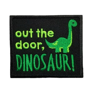 May include: A black embroidered patch with green text that reads "out the door, DINOSAUR!" and a green cartoon dinosaur.
