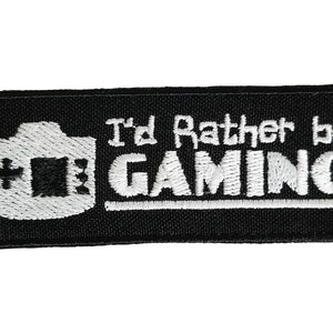 Może przedstawiać: Czarna naszywka haftowana z białym tekstem "I'd Rather Be GAMING" i białą ikoną kontrolera do gier.